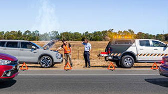 Breakdown service vehicle with flashing lights assisting a car on roadside, warning triangles placed.