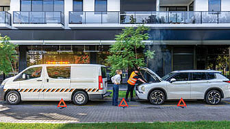 Breakdown service van with flashing lights helping a car on suburban street, warning triangles placed.